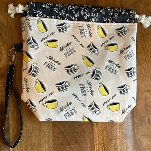 May include: A beige drawstring project bag featuring a repeating pattern of yellow and black coffee mugs and the text "Life's short talk FAST". The bag has a black floral print top section with white drawstrings and a black wrist strap with a metal clip.
