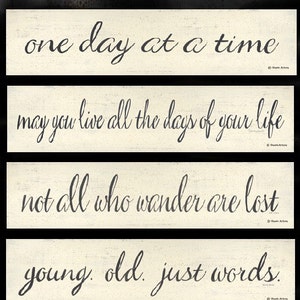 May include: Five white wooden signs with black calligraphy text. The signs read: "one day at a time", "may you live all the days of your life", "not all who wander are lost", "young. old. just words.", and "follow your heart".