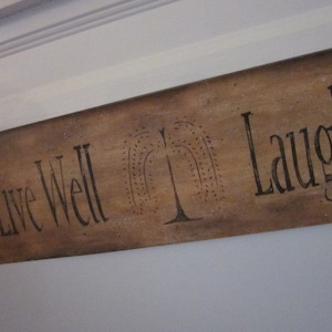 May include: A wooden sign with the text "Live Well Laugh Often" painted in black. The sign has a brown background and a stylized tree design.