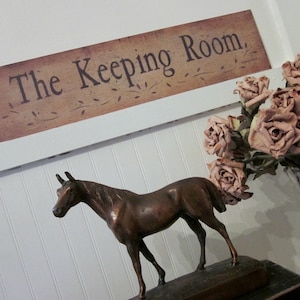 May include: A brown wooden sign with the text "The Keeping Room" in black lettering. A bronze horse figurine is on a wooden base in front of the sign. Dried pink roses are in a vase behind the horse.