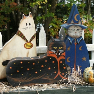 Peut inclure: Trois figurines de chat d'Halloween en bois. Un chat blanc avec un collier noir et une étiquette "BOO", un chat noir avec un collier orange et un chapeau de sorcière, et un chat noir avec une robe bleue et un motif d'étoiles.