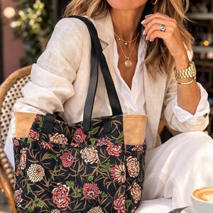 Może przedstawiać: Czarna torba typu tote z kwiatowym wzorem z różowych, białych i zielonych kwiatów. Torba ma czarne uchwyty i beżowe kieszenie boczne. Osoba nosi okulary przeciwsłoneczne, białą koszulę i beżową marynarkę.