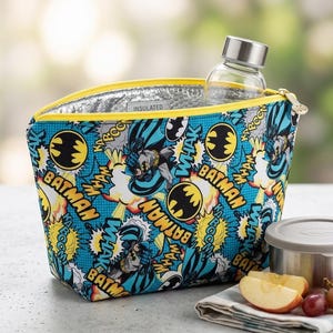 May include: An insulated lunch bag featuring a vibrant blue comic book print with Batman, yellow bat logos, and the text "WHAK" and "KABOOM". The bag has a yellow zipper closure and a silver interior lining, with a clear glass water bottle inside.