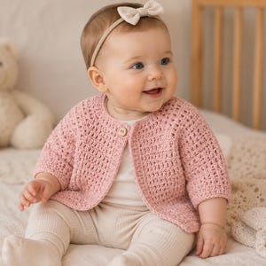 May include: A dusty pink crochet cardigan featuring a round neckline, three-quarter length sleeves, and a single wooden button closure at the top. The garment is crafted with a textured stitch pattern and is worn over a plain white shirt and leggings.