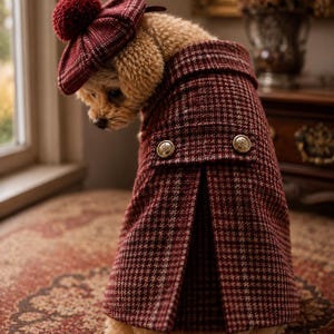 Puede incluir: Un perro pequeño con un abrigo de cuadros en rojo oscuro y beige, botones dorados y una boina a juego con un pompón rojo oscuro. El abrigo tiene un cuello estructurado y un diseño plisado en la parte posterior para un ajuste elegante.
