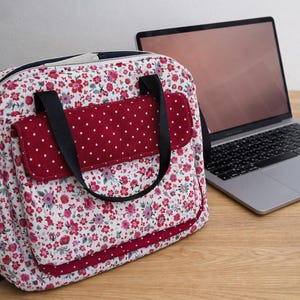 Könnte beinhalten: Eine Tragetasche mit floralem Muster, einer roten Tasche mit Polka-Dots und schwarzen Trägern steht auf einem Holzschreibtisch neben einem Laptop, einer Maus und einem Smartphone. Die Tasche hat einen weißen Hintergrund mit roten und rosa Blumen.