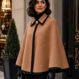 May include: A camel-colored cape featuring a black trim along the collar, front opening, and hem. The garment includes a single gold-toned circular button at the neckline and is worn over a black turtleneck sweater with black leather gloves.