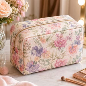 Peut inclure: Une trousse de toilette rectangulaire dotée d'une base crème avec un motif floral dans des tons de rose, violet et vert. La trousse présente une fermeture éclair dorée sur le dessus et une forme structurée avec des coins arrondis.