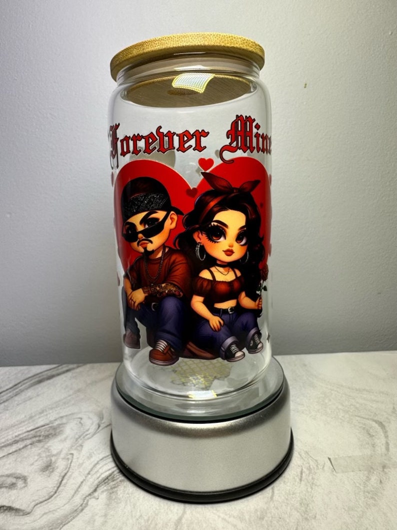 Op de afbeelding: Een glazen pot met een houten deksel en een zilveren voet. De pot heeft een afbeelding van een cartoonkoppel met de tekst "Forever Mine" erop.