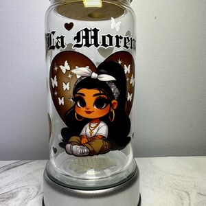 Op de afbeelding: Een glazen pot met een bamboe deksel en een zilveren voet. De pot heeft een cartoon afbeelding van een meisje met bruin haar en bruine ogen, dat een witte bandana en een gouden ketting draagt. De pot heeft de tekst "La Morena" erop.