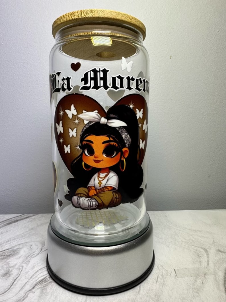 Op de afbeelding: Een glazen pot met een bamboe deksel en een zilveren voet. De pot heeft een cartoon afbeelding van een meisje met bruin haar en bruine ogen, dat een witte bandana en een gouden ketting draagt. De pot heeft de tekst "La Morena" erop.