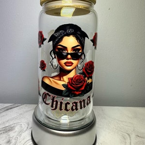 Op de afbeelding: Een glazen pot met een houten deksel en een afbeelding van een vrouw met een zonnebril en een bandana. De pot heeft het woord "Chicana" erop gedrukt en staat op een zilveren voet.