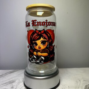 Op de afbeelding: Een glazen pot met een houten deksel en een zilveren voet. De pot heeft een cartoonafbeelding van een meisje met rood haar en een rode bandana. De tekst "La Enojona" is op de pot gedrukt.