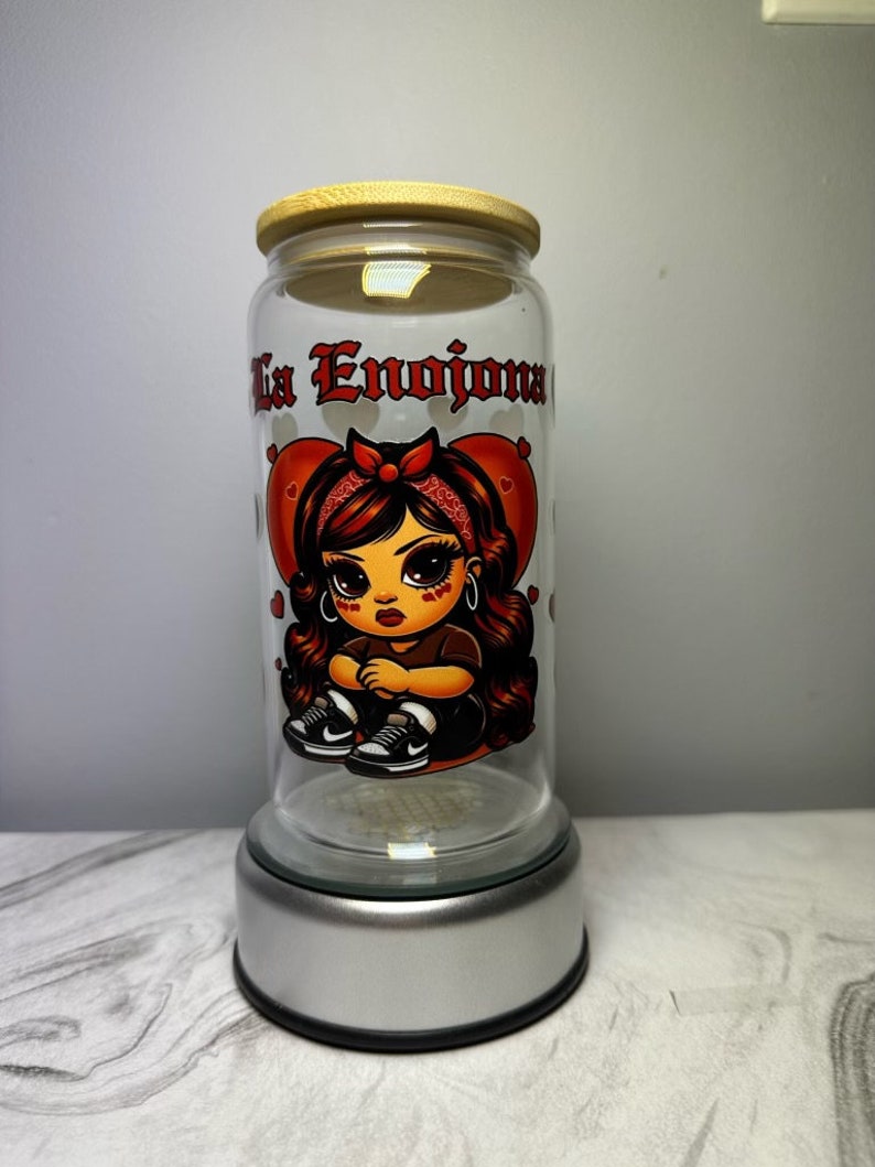 Op de afbeelding: Een glazen pot met een houten deksel en een zilveren voet. De pot heeft een cartoonafbeelding van een meisje met rood haar en een rode bandana. De tekst "La Enojona" is op de pot gedrukt.
