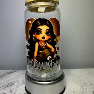 Op de afbeelding: Een glazen pot met een bamboe deksel en een zilveren voet. De pot heeft een cartoon afbeelding van een meisje met lang zwart haar, dat een groene en zwarte outfit draagt, en de woorden "Baby Girl" staan op de voorkant gedrukt.