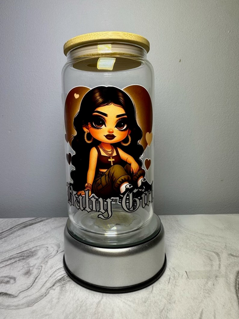 Op de afbeelding: Een glazen pot met een bamboe deksel en een zilveren voet. De pot heeft een cartoon afbeelding van een meisje met lang zwart haar, dat een groene en zwarte outfit draagt, en de woorden "Baby Girl" staan op de voorkant gedrukt.