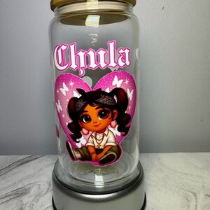 Op de afbeelding: Een glazen pot met een roze hartontwerp met een cartoonmeisje met een roze bandana en een gouden ketting. De pot heeft een houten deksel en een zilveren basis. De pot is gelabeld met "Chula".