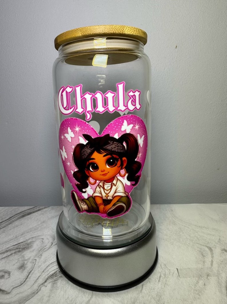 Op de afbeelding: Een glazen pot met een roze hartontwerp met een cartoonmeisje met een roze bandana en een gouden ketting. De pot heeft een houten deksel en een zilveren basis. De pot is gelabeld met "Chula".
