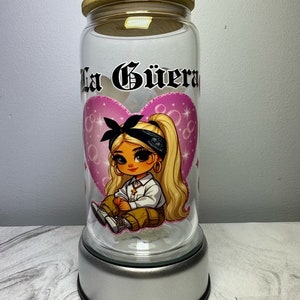 Op de afbeelding: Een glazen pot met een houten deksel en een zilveren voet. De pot heeft een roze hart met een cartoonmeisje dat een bandana en een wit shirt draagt. De tekst "La G&uuml;era" is op de pot gedrukt.