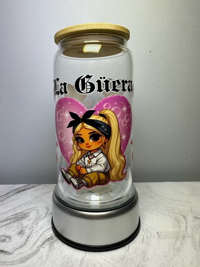 Op de afbeelding: Een glazen pot met een houten deksel en een zilveren voet. De pot heeft een roze hart met een cartoonmeisje dat een bandana en een wit shirt draagt. De tekst "La G&uuml;era" is op de pot gedrukt.