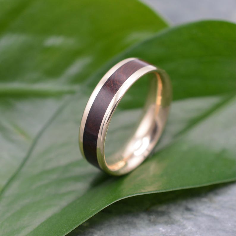 May include: A gold ring with a dark brown wood inlay. The ring is set against a green leaf, highlighting the contrast between the warm gold and the rich wood grain. The ring's band is smooth and polished.