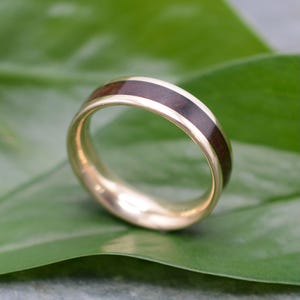 May include: A gold ring with a dark brown wood inlay. The ring is set against a green leaf, highlighting the contrast between the warm gold and the rich wood grain. The band is approximately 0.25 inches wide.