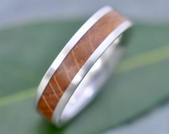 May include: A silver ring with a wooden inlay. The ring features a band of light brown wood encased in a polished silver setting. The wood grain is visible, adding texture to the design. The ring is a simple, elegant piece of jewelry.