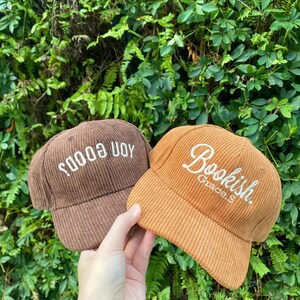 May include: Two corduroy baseball caps. One is brown with the text "soods uoy" in white. The other is orange with the text "Bookish. Grace.S" in white. The caps are held in front of a background of green foliage.