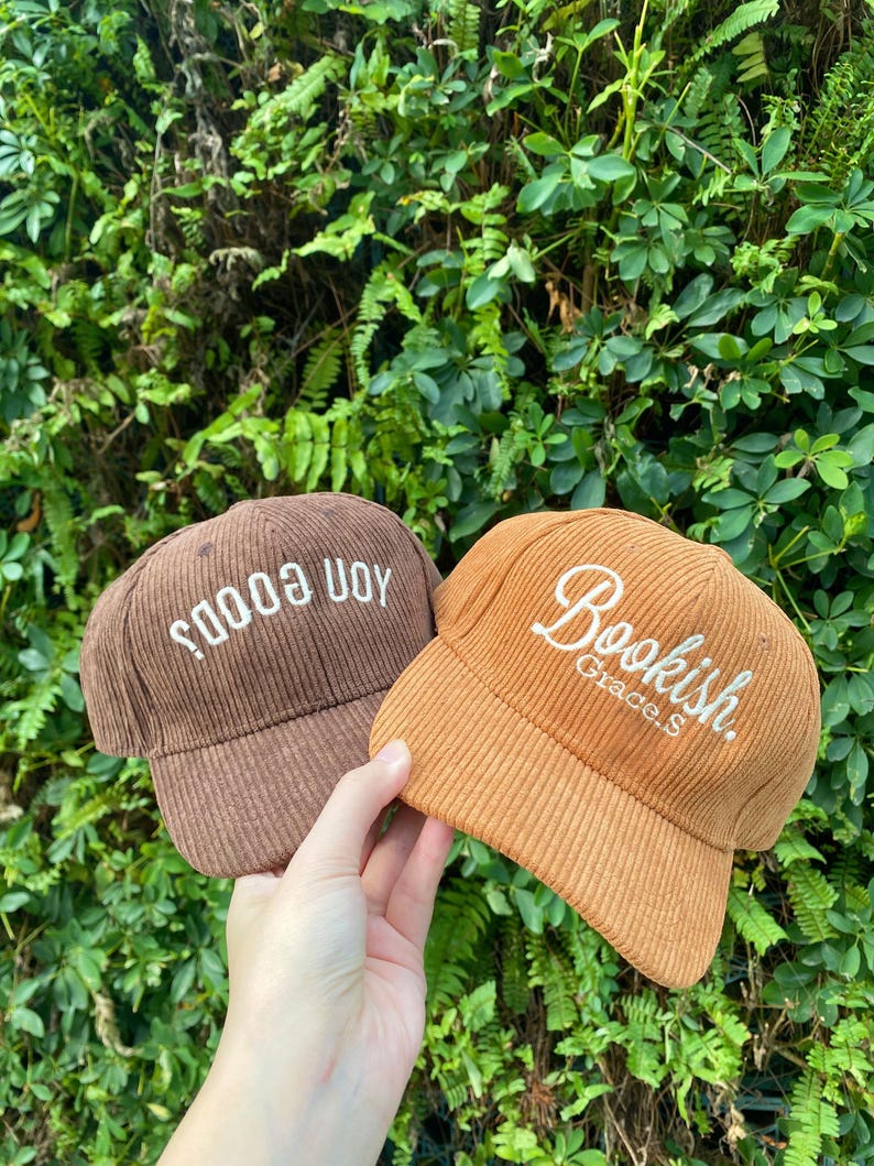 May include: Two corduroy baseball caps. One is brown with the text "soods uoy" in white. The other is orange with the text "Bookish. Grace.S" in white. The caps are held in front of a background of green foliage.