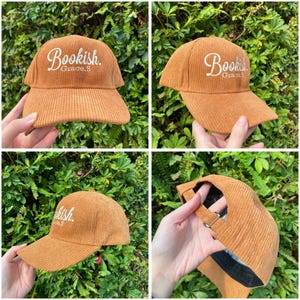 May include: A corduroy baseball cap in a warm brown color. The cap features the word "Bookish" embroidered in white script on the front. The brim is curved, and the cap has an adjustable strap.