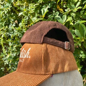 May include: Three corduroy baseball caps in shades of brown, tan, and beige are stacked. The middle cap has the word "kish" embroidered in white. Each cap has a curved brim and an adjustable strap.