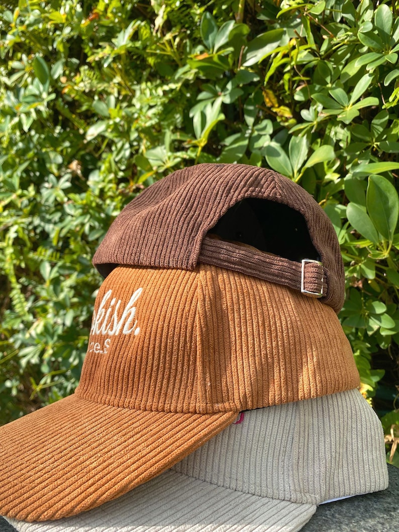 May include: Three corduroy baseball caps in shades of brown, tan, and beige are stacked. The middle cap has the word "kish" embroidered in white. Each cap has a curved brim and an adjustable strap.