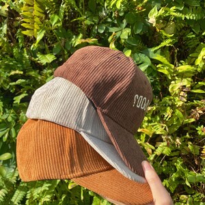 May include: Three corduroy baseball caps in shades of brown, tan, and orange are stacked together. The top cap has the word "SOOD" embroidered on the side. The caps are held in a hand against a backdrop of green foliage.