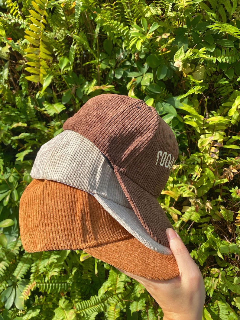 May include: Three corduroy baseball caps in shades of brown, tan, and orange are stacked together. The top cap has the word "SOOD" embroidered on the side. The caps are held in a hand against a backdrop of green foliage.