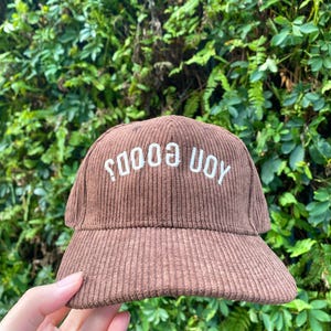 May include: A brown corduroy baseball cap with the text "SOOOO UOY" embroidered in white. The cap has a curved brim and a structured crown. The background is a lush green foliage.
