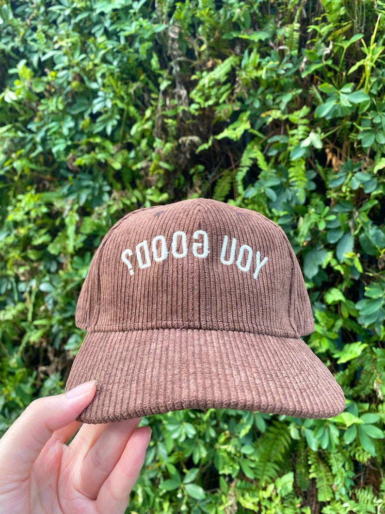 May include: A brown corduroy baseball cap with the text "SOOOO UOY" embroidered in white. The cap has a curved brim and a structured crown. The background is a lush green foliage.