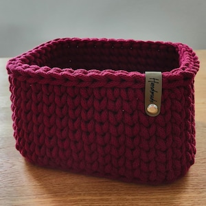 Könnte beinhalten: Ein rechteckiger Häkelkorb in Burgunderrot. Der Korb ist aus dickem Garn gefertigt und hat ein strukturiertes Stichmuster. Ein kleines braunes Lederetikett mit dem Wort "Handmade" ist mit einem silbernen Knopf befestigt.