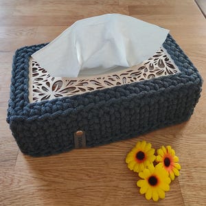 Könnte beinhalten: Handgefertigte, dunkelgraue Taschentuchbox-Abdeckung mit dekorativem Holzeinsatz. Die Abdeckung ist mit dickem Garn gehäkelt und hat ein kleines Lederetikett. Drei gelbe Sonnenblumen mit orangefarbenen Zentren liegen neben der Box.