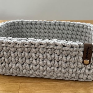 Könnte beinhalten: Ein rechteckiger, hellgrauer Häkelkorb mit einem braunen Lederanhänger mit der Aufschrift "Handmade". Der Korb besteht aus dickem, strukturiertem Garn und steht auf einer Holzoberfläche. Der Korb ist etwa 25 cm lang, 15 cm breit und 10 cm hoch.
