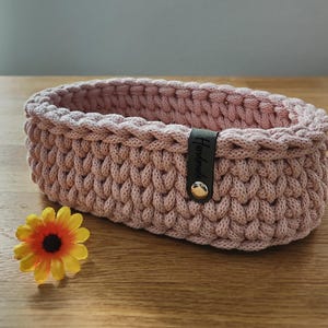 Könnte beinhalten: Ein rechteckiger Häkelkorb in Staubrosa mit einem dunklen Lederetikett mit der Aufschrift "Handmade". Der Korb ist aus dickem, strukturiertem Garn gefertigt und hat einen Knopfverschluss. Eine gelb-orangefarbene Blume liegt neben dem Korb.
