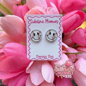 May include: A pair of baseball-themed stud earrings with a smiling face design. The earrings are on a white card with the text "Sideline Mamas" and "Stainless Steel". The background features pink flowers.