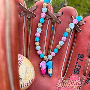 May include: A colorful necklace with a pendant featuring the number "11" in an ice cream cone design, resting on a baseball glove. The necklace has pink, blue, and gold beads. A baseball is also visible.