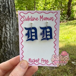 Może przedstawiać: Niebieskie i białe kolczyki z literą "D" na r&oacute;żowej karcie z napisem "Sideline Mamas" i "Nickel Free". Kolczyki wiszą na srebrnych sztyftach. Tło to naturalne zielone otoczenie.