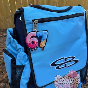 May include: A light blue backpack with black trim and zippers. A pink and blue number 67 ice cream charm hangs from the zipper. The backpack has a logo that says "Sideline Mamas".