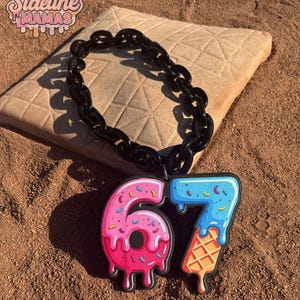 May include: A black chain necklace with a pendant featuring the number "67" in a playful, colorful design. The "6" is styled as a pink donut with sprinkles, and the "7" as a blue ice cream cone. The text "Sideline Mamas" is in the upper left corner.