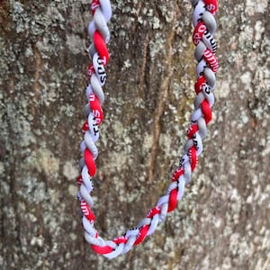 May include: Braided necklace in red, white, and gray. The necklace is made of interwoven fabric strands. The red strands have white text. The necklace is displayed against a textured, brown background.