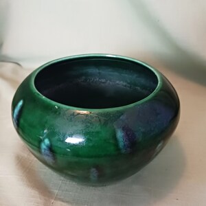 May include: A round, ceramic bowl with a glossy, emerald green glaze. The bowl has a wide opening and a rounded shape. Darker blue-green accents are visible on the surface. The bowl is likely used for decorative purposes or as a small planter.