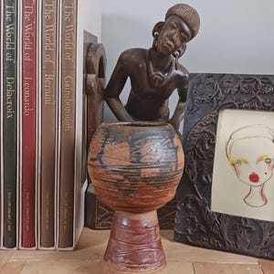 May include: A handmade ceramic vase with a bulbous top and a pedestal base. The vase features brown and black abstract patterns. The background includes art books, a wooden sculpture, and a framed drawing.