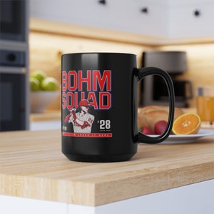 May include: Black ceramic coffee mug with a white graphic of a baseball player in a red and white uniform. The text "BOHM SQUAD" is in red and blue letters above the player. The text "#28 THIRD BASE" is in red and white letters below the player. The text "WE ALL JUST IN LOVES IT HERE" is in white letters below the player.