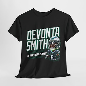 Puede incluir: Camiseta negra con un gráfico blanco del receptor abierto de los Philadelphia Eagles DeVonta Smith vistiendo su camiseta y casco. El texto "DeVonta Smith #6 The Slim Reaper" está impreso en la camiseta.